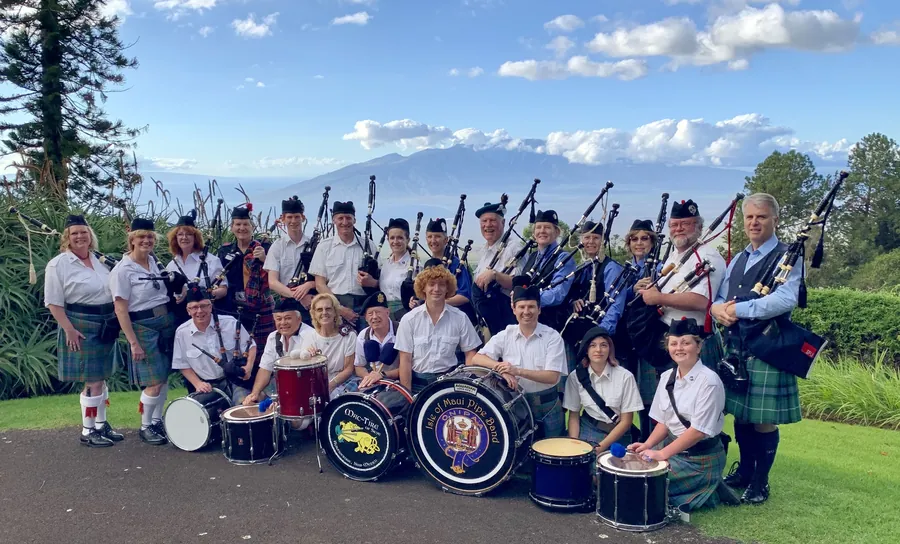 The full band in kilts with drums on a lawn, Maui mountains behind