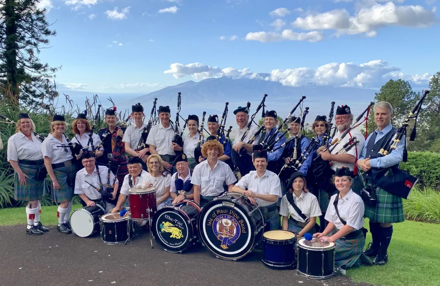 The full band in kilts with drums, posed on a lawn with Maui mountains behind